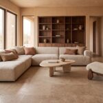 Warm minimalist living room with a cream corner sofa, pale sculptural coffee table, dark walnut shelving and sunlit clay walls.