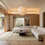 Warm Japandi living room with cream sectional sofa, dark wood coffee table, tatami floor, paper lantern and shoji doors overlooking a courtyard